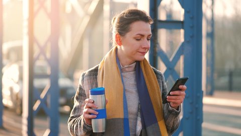 Portrait of a young caucasian businesswoman in a coat, walking around the city on a frosty morning, drinking coffee and using smartphone. Communication, work day, busy life concept. Slow motion