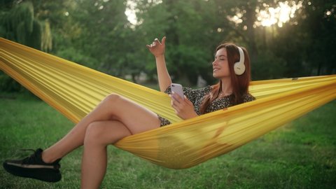 Young Cheerful red-haired woman resting on hammock. Pretty lady enjoys listening chill music. Beautiful girl wear headphones having fun