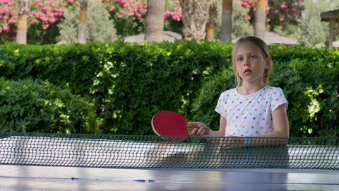 Girl child hit lightweight ball, play table tennis, known as ping pong. Hard table divided by tennis net use small rackets. Real people, leisure game. Positive emotion