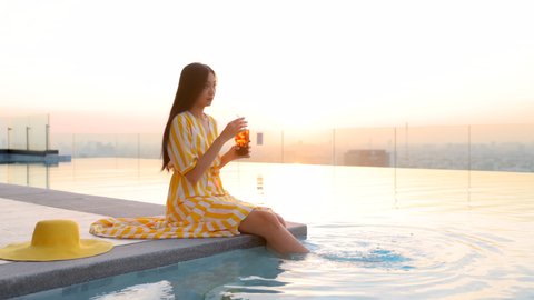 Easy relax asian businesswoman lifestyle in beautiful summer dress sitting on the ledge of the pool and enjoying refreshing cocktail in a glass with ice on Blurred background beautiful sunset sky.