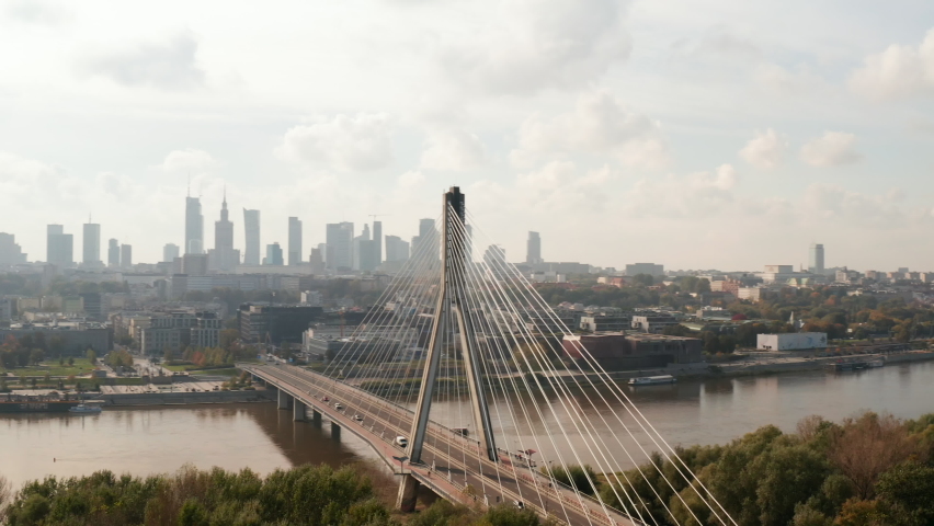 Slide and pan shot of bridge over Vistula river with reversed Y shape pillar. Cityscape against bright sky in background. Warsaw, Poland