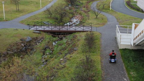 Aerial view of a caucasian man with amputated both legs and both arms driving on electric wheelchair along the empty path and over small bridge in park. Autumn evening