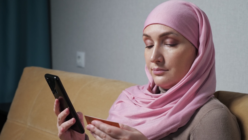Pregnant Muslim woman wearing hijab types phone number from business card into smartphone sitting on comfortable sofa in living room closeup.