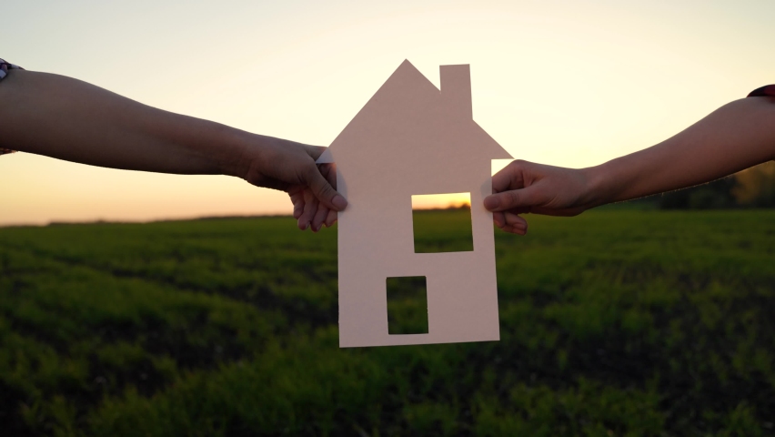 Home and happiness symbol. Family holds paper house at sunset, sun shines through window. Camera movement to window. Concept of building house for family. My dream is to build, buy house for children