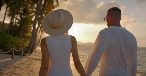 Back view man and woman in hat walking hand in hand along the ocean sandy beach enjoying their date 