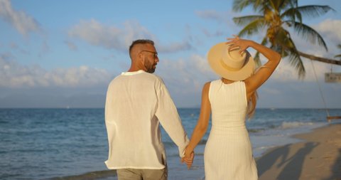 Young couple in white clothes enjoying tropical sunset on the sandy beach by walking hand in hand 