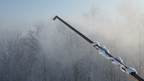 Snow gun making artificial snow in morning, on back fir forest. Snowmaking by water through snow cannon for snow cover ski slopes in ski resort. Snowfall from snowgun in sun. No people. 4k.