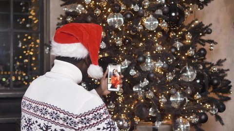 African-American man in a Christmas hat communicates via video calls on a smartphone online with his girlfriend against the background of a Christmas tree