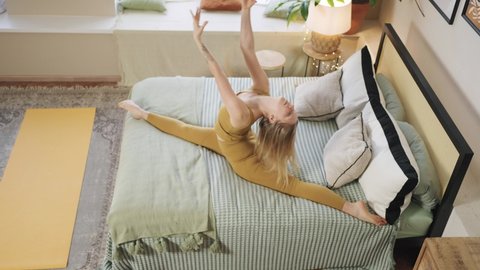 Young blonde-haired woman wearing sporty yellow top and leggings does split stretching on bed in spacious bedroom upper view