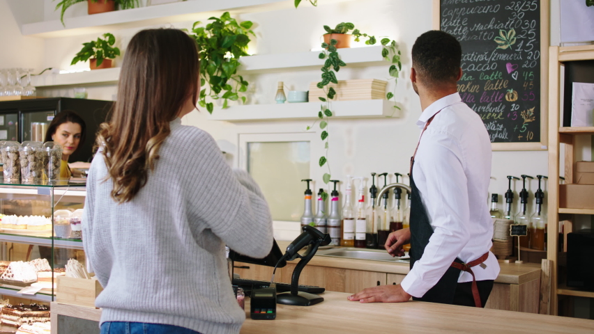 In front of the camera in the cozy coffee shop Afro American guy barista working at the terminal desk he discussing with customer lady and take the order