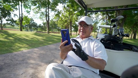 Asian senior businessman sitting on golf cart with using mobile phone working online corporate business while golfing with at country club. Elderly male golfer enjoy outdoor sport in summer vacation.