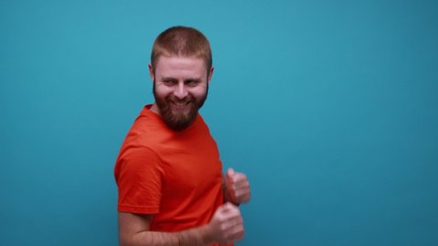 Positive full of energy bearded man dancing to the music, having fun, resting and enjoying life, party, entertainment, wearing orange T-shirt. Indoor studio shot isolated on blue background.