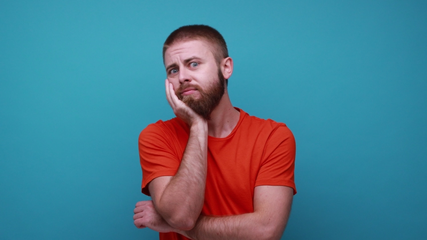 Thoughtful bearded man holding his chin and pondering idea, confused not sure about solution, having pensive expression, wearing orange T-shirt. Indoor studio shot isolated on blue background. Royalty-Free Stock Footage #1083969619