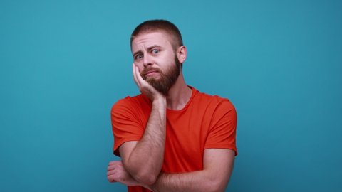 Thoughtful bearded man holding his chin and pondering idea, confused not sure about solution, having pensive expression, wearing orange T-shirt. Indoor studio shot isolated on blue background.