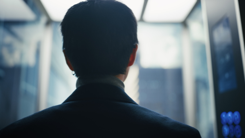 Stylish Japanese Businessman Riding Glass Elevator to Office in Modern Business Center. Male Looking at Modern Downtown Skyscrapers Out of the Panorama Window in the Lift. Back Turned to Camera. | Shutterstock HD Video #1083971905