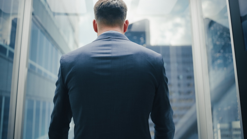 Portrait of a Successful Businessman in a Suit Riding Glass Elevator to Office in Modern Business Center. Male Turning Around to the Camera, Crossing Arms, Charmingly Smiling and Striking a Pose. Royalty-Free Stock Footage #1083972001