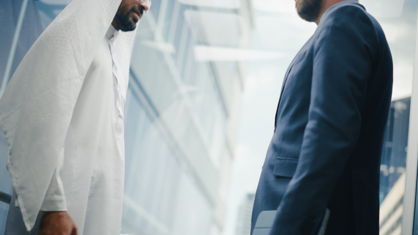 Businessman Talking with Arab Investment Partner while Riding Glass Elevator to Office in a Modern Business Center. International Corporate Associates Shake Hands and Agree on a Deal in a Lift. Royalty-Free Stock Footage #1083972220