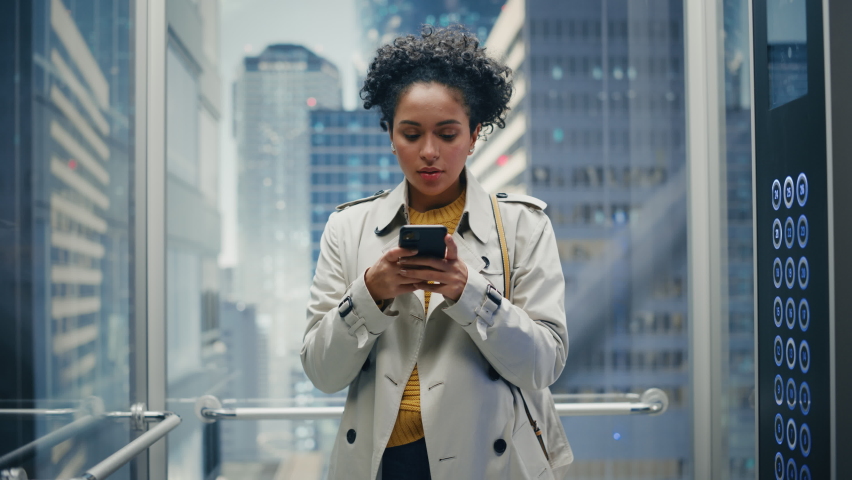 Beautiful Black Female Riding Glass Elevator to Office in Modern Business Center. Successful Manager Smile while Using Smartphone, Write Text Message, Check Social Media and Work Emails in a Lift. | Shutterstock HD Video #1083972268