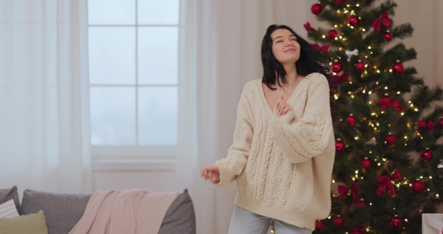 Funny girl dancing, having fun while celebrating winter holidays. A happy festive woman celebrating New Year or Xmas near decorated Christmas tree indoors