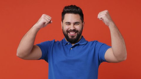 Excited jubilant overjoyed happy young bearded brunet man 20s years old wear blue t-shirt doing winner gesture celebrate clenching fists say yes isolated on plain red orange background studio portrait
