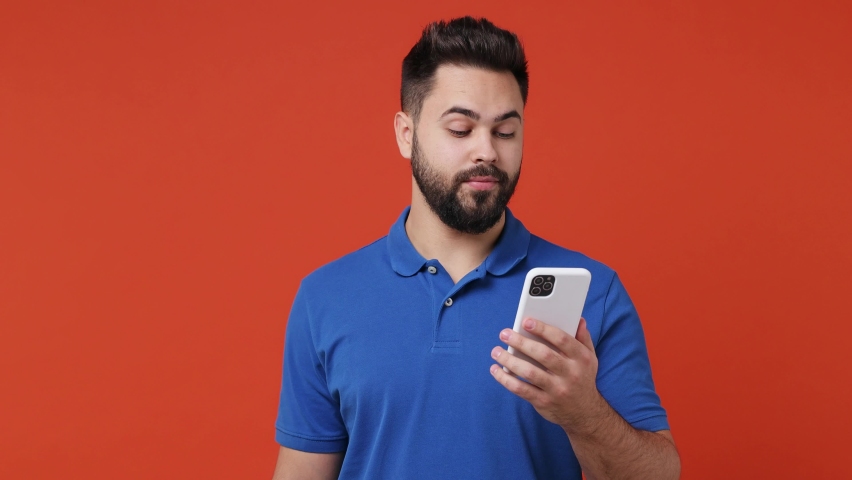 Happy calm smiling young bearded brunet man 20s years old wears blue t-shirt hold use talk on mobile cell phone conducting pleasant conversation isolated on plain red orange background studio portrait