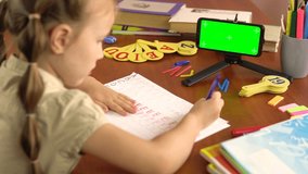 Little schoolgirl writes latin letters sitting at the table with a smartphone with a green screen.4k