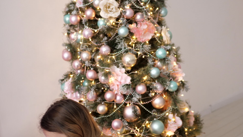 mom and daughter in a pink dress and wrapped in a garland are sitting next to the Christmas tree at home