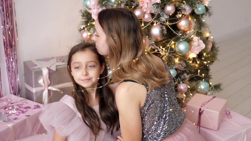 mom and daughter in a pink dress and wrapped in a garland are sitting next to the Christmas tree at home