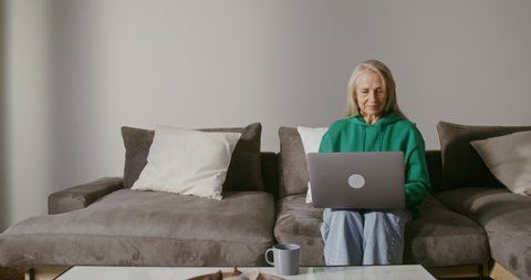 Elderly gray-haired woman of European appearance, dressed in casual clothes, uses laptop while sitting on sofa in modern home interior