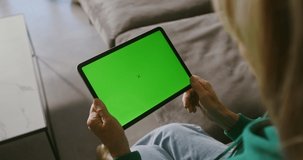 An elderly woman is video chatting on a tablet with a green screen, holding it in front of her. Copy space, chroma key, woman's face is not visible, video filming from behind