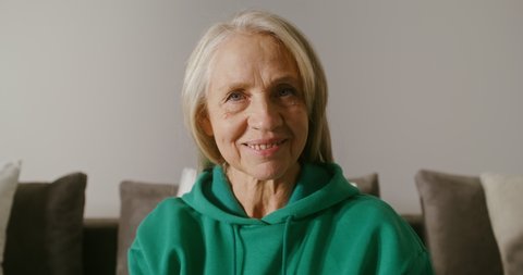 Elderly woman with long gray hair, dressed in casual clothes, smiling while looking at the camera, sitting on the sofa, close-up