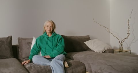 A beautiful elderly woman of European appearance with long gray hair is sitting on a sofa in a modern home interior, her leg bent under her in a shoe and smiling while looking at the camera