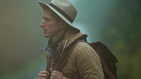 close up portrait of man hitchhiker wearing in retro style. traveler in hat with backpack stands outdoors at road in cold wet foggy autumn day and waites a car. soft focus