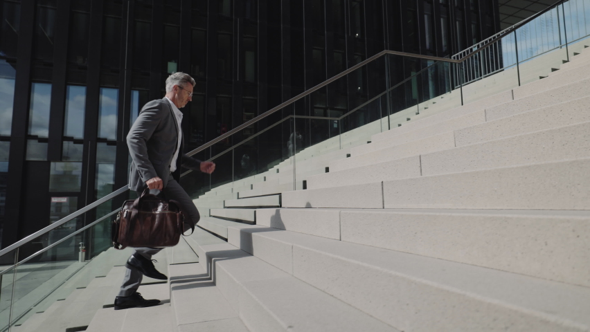 Side view of caucasian aged man in stylish suit and eyeglasses walking up on stairs with suitcase in hands. Busy male emloyer in hurry. | Shutterstock HD Video #1083992986