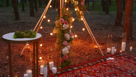 Bohemian tipi wooden arch decorated with burning candles, roses and pampass grass, wrapped in fairy lights illumination on outdoor wedding ceremony venue in pine forest at night. Bulbs garland shines.