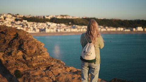Tourist traveler woman with backpack stands on rocks of Nazare town. European tourism, traveling in Portugal. Mountains of Forte de Sao Miguel Arcanjo. Tourists take phone pictures, film sea or ocean.