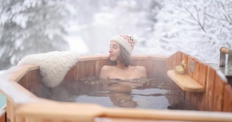 Woman relaxing in hot bath outdoors, enjoying thermal spa at snowy mountains. Winter holidays in the mountains, hot water treatments concept. Caucasian woman wearing winter hat