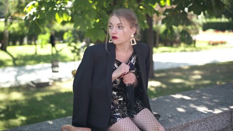Young female drinker looking around sitting in urban park drinking wine from bottle. Portrait of slim beautiful Caucasian millennial woman in dress and jacket after all night partying outdoors
