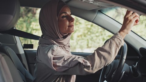 Beautiful Muslim woman in hijab getting in car, putting seat belt on and looking in mirror with smile before driving