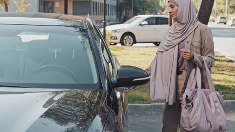Muslim woman in hijab and casualwear walking to car with gym bag and getting inside before driving