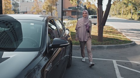 Muslim woman in hijab and casual outfit walking on parking, getting in car and sitting on driver seat