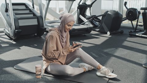 Portrait of beautiful Muslim woman in hijab and sportswear sitting on exercise mat in gym, using smartphone an then looking at camera and smiling
