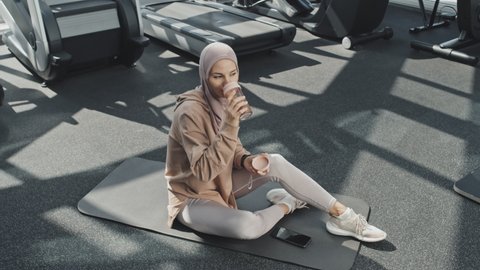 High angle shot of Muslim woman in hijab sitting on exercise mat on floor in gym and drinking water from bottle