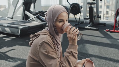 Beautiful Muslim woman in hijab sitting on floor in gym and drinking water from shaker bottle