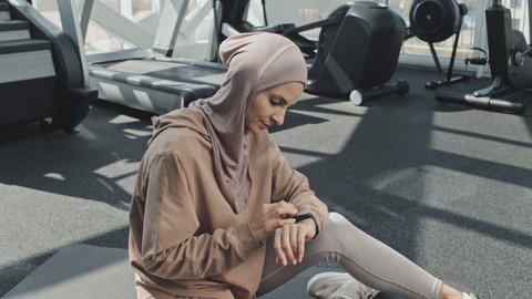 Zoom in shot of sporty Muslim woman in hijab sitting on mat in gym and setting fitness app on smartwatch