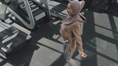 High angle tilt up shot of Muslim woman in hijab and sportswear jumping the rope in gym