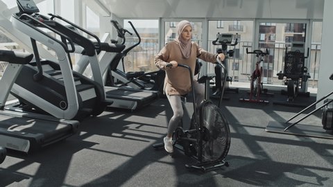 Muslim woman in hijab and sportswear riding stationary exercise bike while training at gym