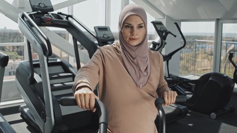 Beautiful Muslim woman in hijab exercising on stationery bike during cycling workout at gym