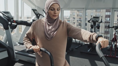 Tilt up shot of young Muslim woman in hijab and sportswear riding stationery bike while training at gym