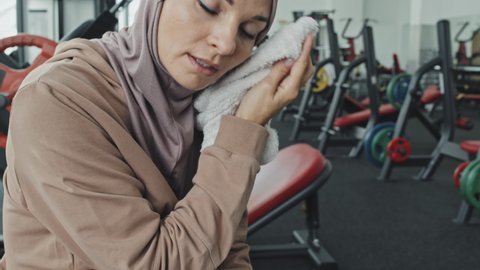 Tilt down shot of beautiful Muslim woman in hijab and sportswear wiping forehead with towel and using digital tablet while resting after workout in gym